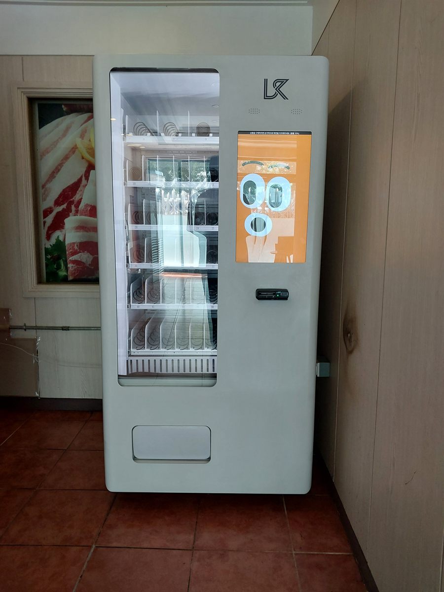 식당 대기 공간 벽면에 세운 냉장 음료 자판기 — 왼쪽에 액자가 걸린 목재 벽, 바닥은 붉은 타일, 오른쪽에 안쪽으로 이어지는 문이 보이는 구성