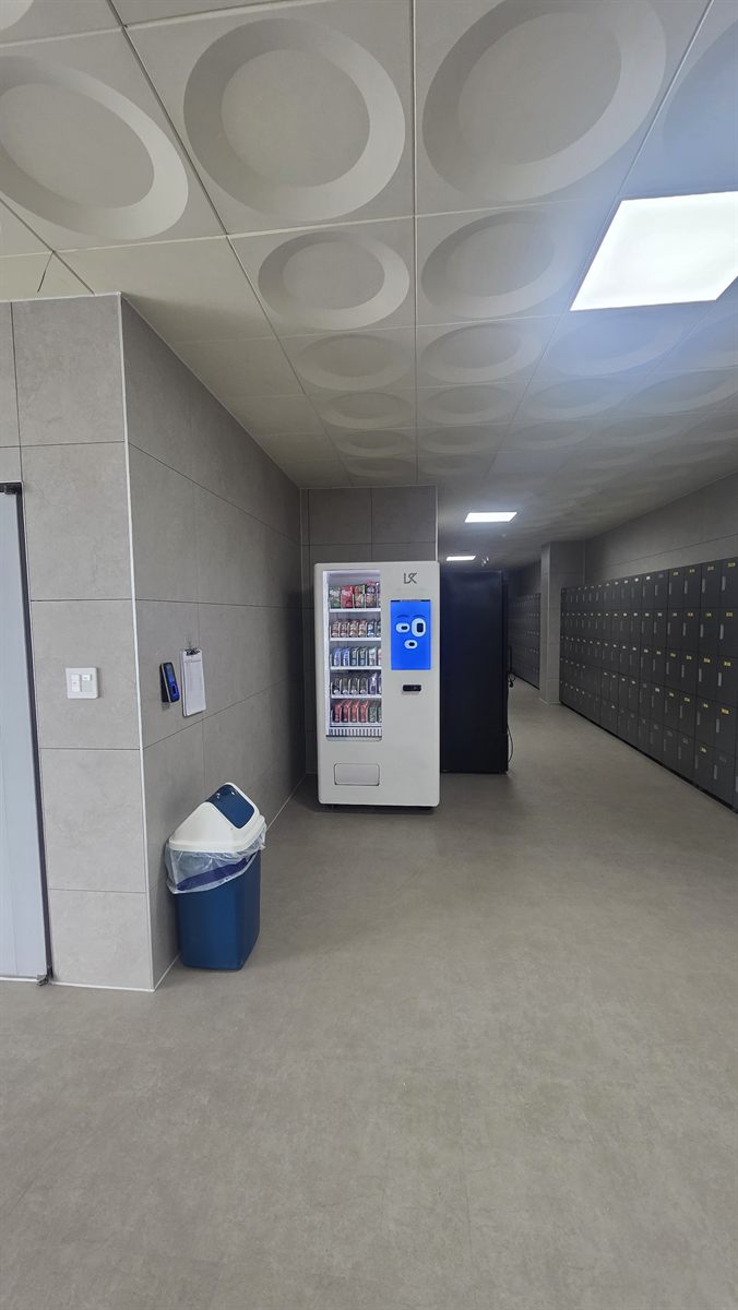 회색 타일 벽과 원형 무늬 천장이 있는 넓은 복도. 오른쪽으로 짙은 회색 사물함이 길게 늘어서 있고, 정면 벽 모서리에 파란 터치화면이 달린 흰색 스마트자판기와 검은 기계 한 대가 서 있으며, 왼쪽 벽 앞에는 파란 휴지통이 놓여 있음